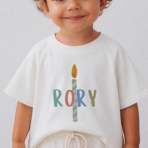 Pode incluir: T-shirt branca de manga curta com um gr&aacute;fico de vela colorida e o nome "RORY" impresso abaixo. A vela tem uma chama amarela e um corpo listrado azul e branco. O nome est&aacute; em v&aacute;rias cores.