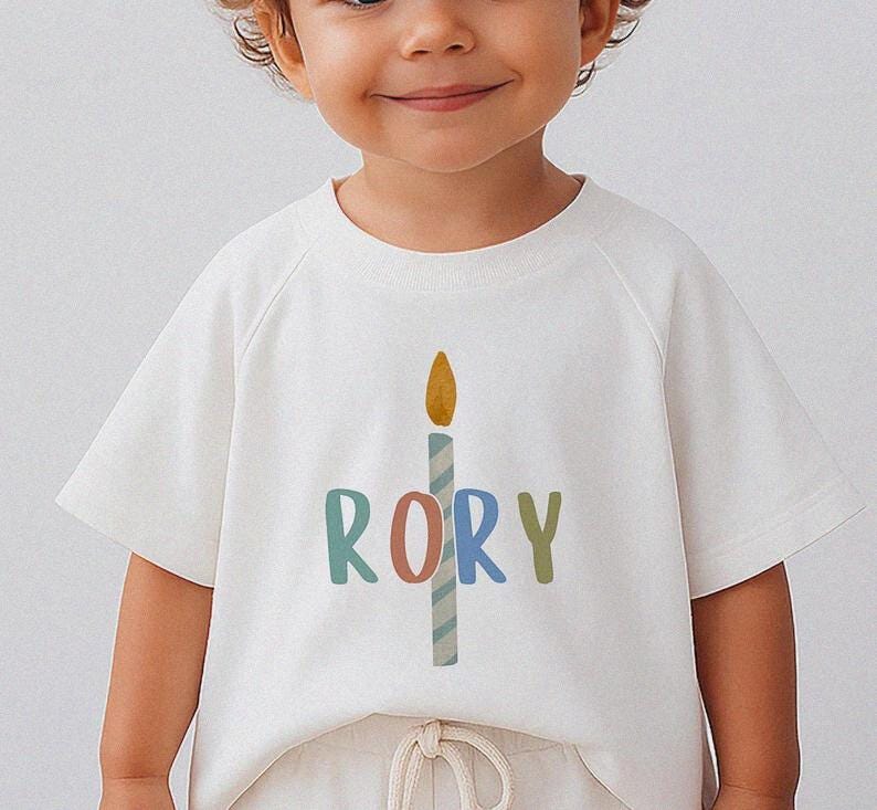 Pode incluir: T-shirt branca de manga curta com um gr&aacute;fico de vela colorida e o nome "RORY" impresso abaixo. A vela tem uma chama amarela e um corpo listrado azul e branco. O nome est&aacute; em v&aacute;rias cores.