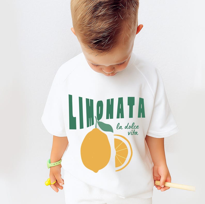 May include: White short-sleeved t-shirt with a green and yellow graphic design. The design features the word "LIMONATA" in green, a yellow lemon, and the phrase "la dolce vita". A fun, casual top.