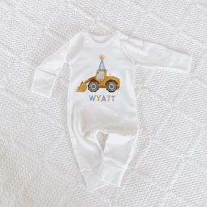 May include: White long-sleeved baby onesie with a yellow construction vehicle wearing a blue party hat with the number one on it. The onesie has the name Wyatt printed below the vehicle.