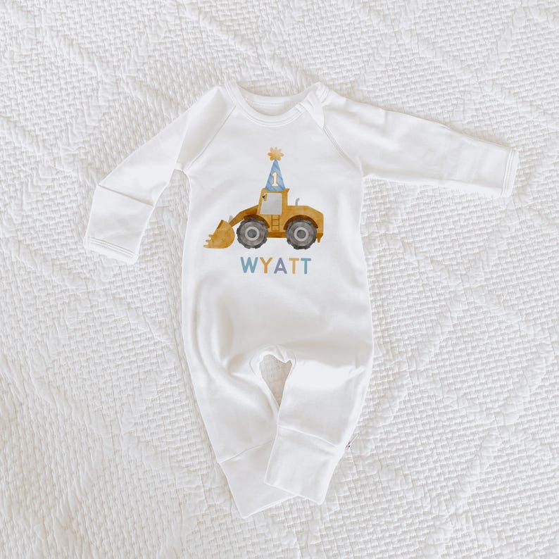 May include: White long-sleeved baby onesie with a yellow construction vehicle wearing a blue party hat with the number one on it. The onesie has the name Wyatt printed below the vehicle.
