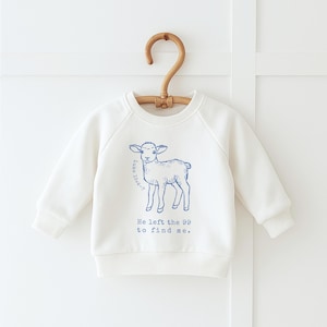 Op de afbeelding: Crèmekleurige baby-sweatshirt met een blauwe illustratie van een lam en de tekst "He left the 99 to find me." De sweatshirt hangt aan een houten hanger.