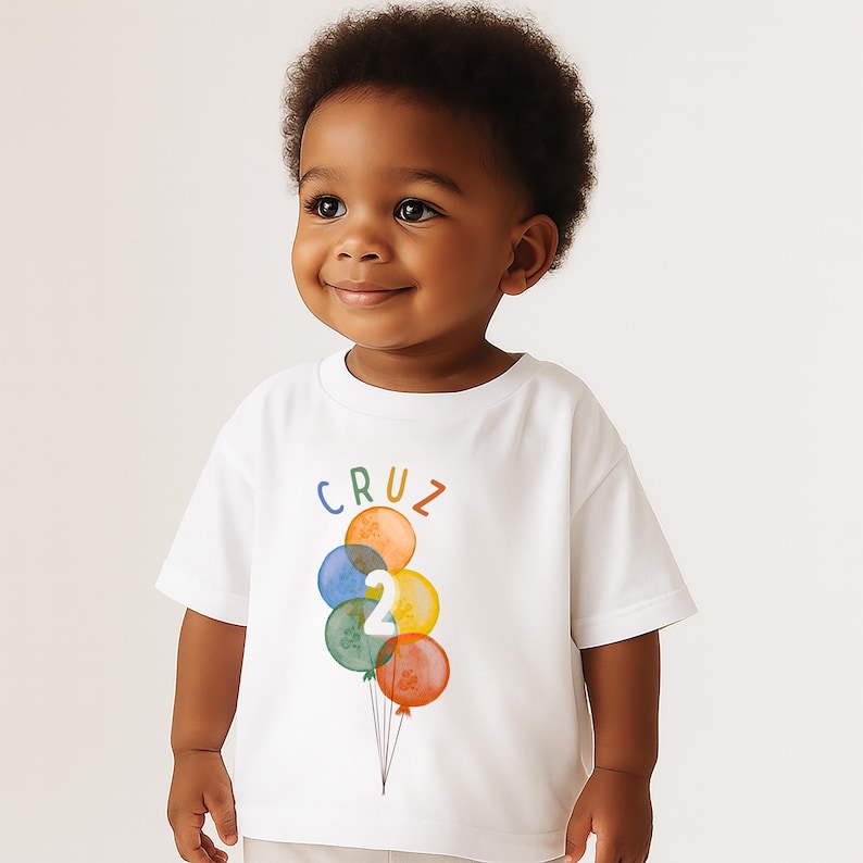 May include: A white t-shirt featuring a colorful balloon design with the name "CRUZ" above and the number "2" on the balloons. The balloons are in various colors, including orange, blue, green, and yellow. The shirt is worn by a young child.