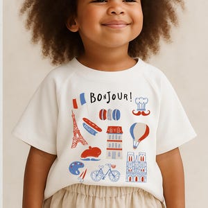 Op de afbeelding: Een wit T-shirt met een Frans thema, met het woord "Bonjour!" en illustraties van de Eiffeltoren, macarons, een baguette en een heteluchtballon. Het shirt heeft korte mouwen en een ronde hals.