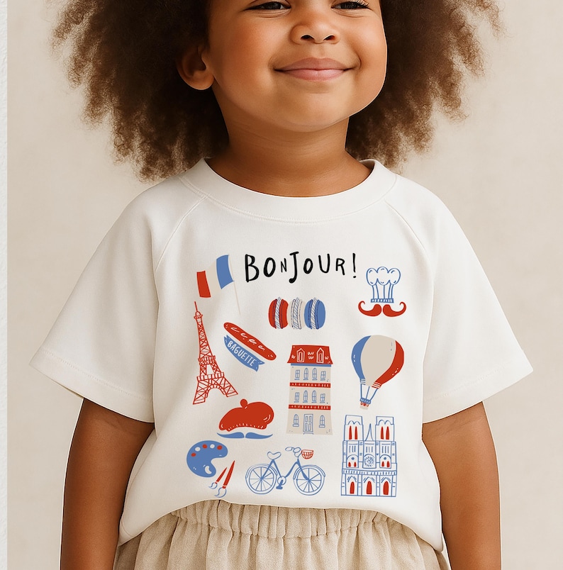 Op de afbeelding: Een wit T-shirt met een Frans thema, met het woord "Bonjour!" en illustraties van de Eiffeltoren, macarons, een baguette en een heteluchtballon. Het shirt heeft korte mouwen en een ronde hals.