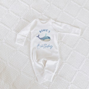 May include: White baby onesie with a blue whale wearing a party hat and the text "BOWIE'S Birthday" on the front.
