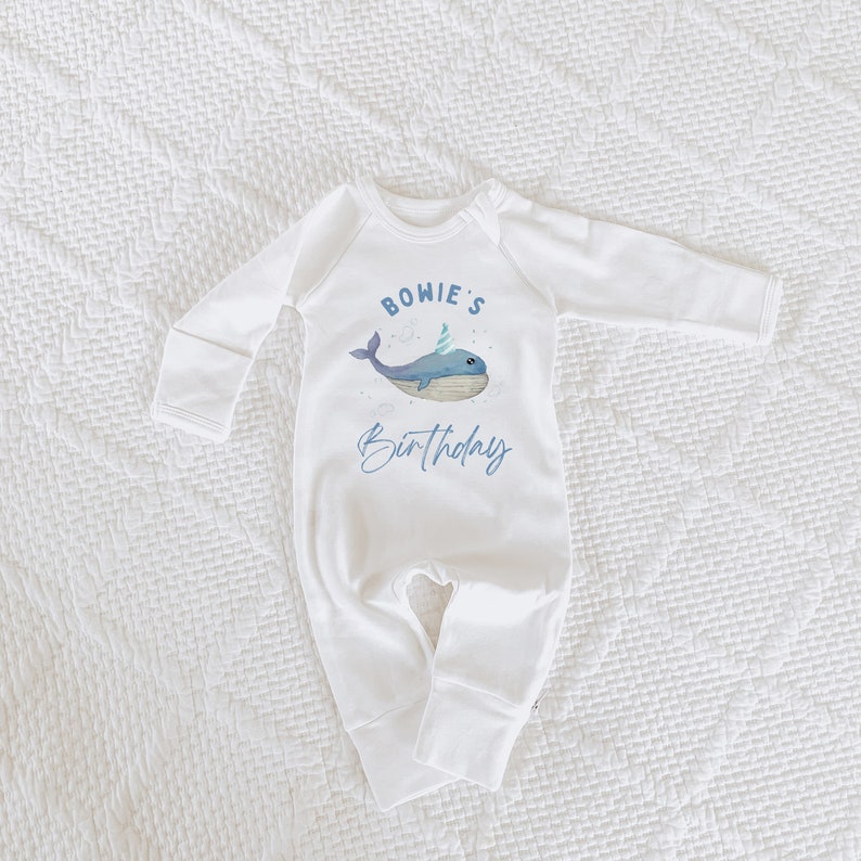 May include: White baby onesie with a blue whale wearing a party hat and the text "BOWIE'S Birthday" on the front.