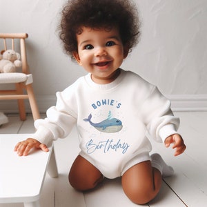 May include: White baby onesie with a blue whale wearing a party hat and the text "BOWIE'S Birthday".