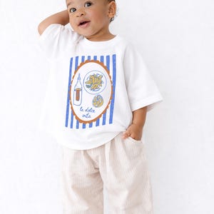 May include: White t-shirt with a blue and brown graphic of food and a bottle, with the text "la dolce vita". The shirt is paired with beige striped pants. The child is smiling and looking up.