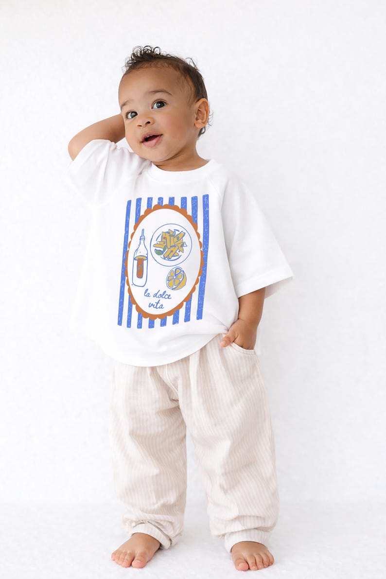 May include: White t-shirt with a blue and brown graphic of food and a bottle, with the text "la dolce vita". The shirt is paired with beige striped pants. The child is smiling and looking up.