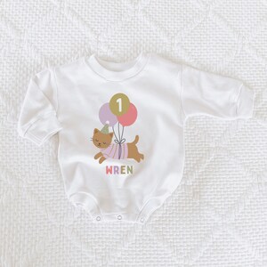 May include: White baby bodysuit with a cartoon cat graphic holding balloons. The cat wears a party hat and a striped shirt. The number "1" is on a balloon. The name "WREN" is printed below the cat in pink.