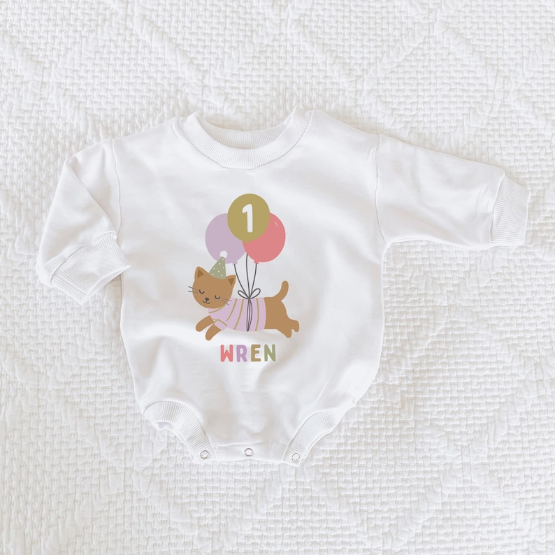 May include: White baby bodysuit with a cartoon cat graphic holding balloons. The cat wears a party hat and a striped shirt. The number "1" is on a balloon. The name "WREN" is printed below the cat in pink.