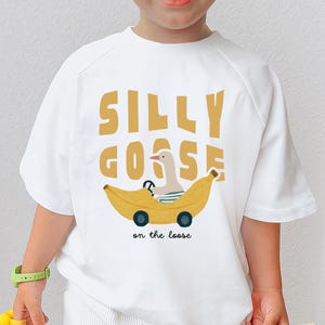 以下が含まれることがあります： 白のTシャツ。バナナの車を運転するガチョウの気まぐれなデザイン。イラストの上には「SILLY GOOSE」、下には「on the loose」と書かれています。カジュアルな半袖スタイルです。