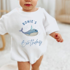 May include: White baby onesie with a blue whale wearing a party hat and the text "BOWIE'S Birthday" in blue.