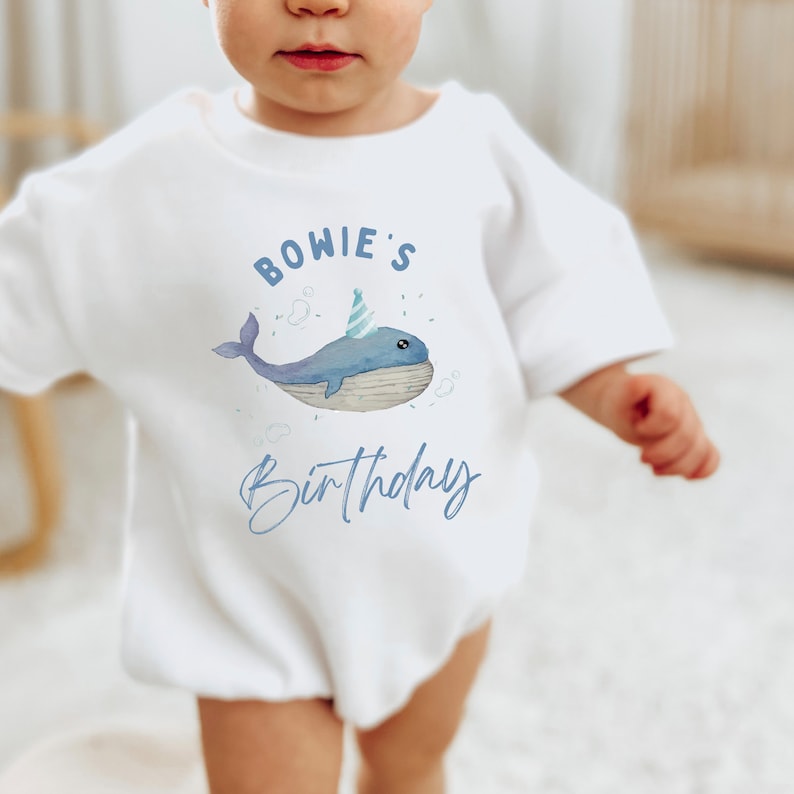 May include: White baby onesie with a blue whale wearing a party hat and the text "BOWIE'S Birthday" in blue.