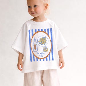 May include: White t-shirt with blue vertical stripes and a brown oval design featuring a bottle, pasta, and lemon illustration. The text "la dolce vita" is below the design. The shirt is worn with pink striped pants.
