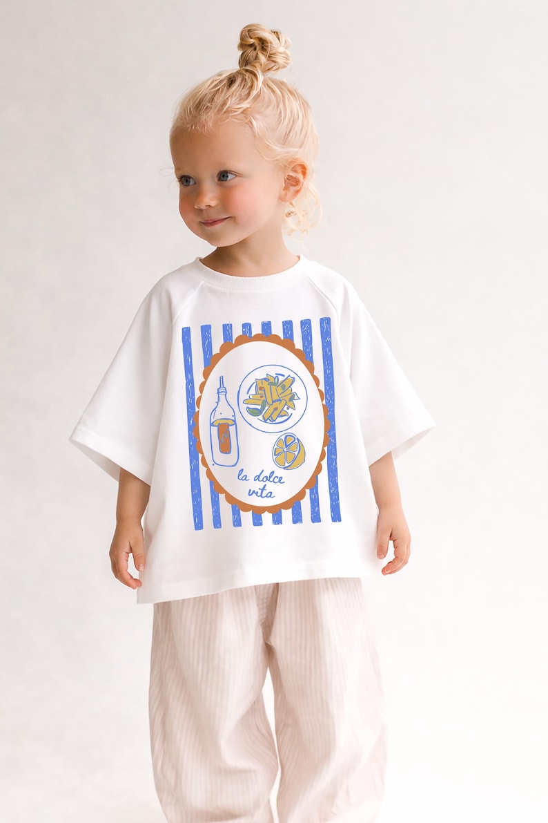 May include: White t-shirt with blue vertical stripes and a brown oval design featuring a bottle, pasta, and lemon illustration. The text "la dolce vita" is below the design. The shirt is worn with pink striped pants.