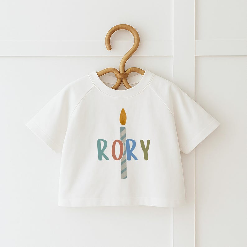 Pode incluir: T-shirt branca de manga curta com um gr&aacute;fico de vela colorida e o nome "RORY" em letras azuis, rosa e verdes. A vela tem uma chama amarela. A camisa est&aacute; pendurada em um cabide de madeira.