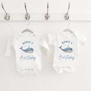 May include: Two white baby onesies with long and short sleeves. The onesies feature a blue whale with a party hat and the text "BOWIE'S Birthday".
