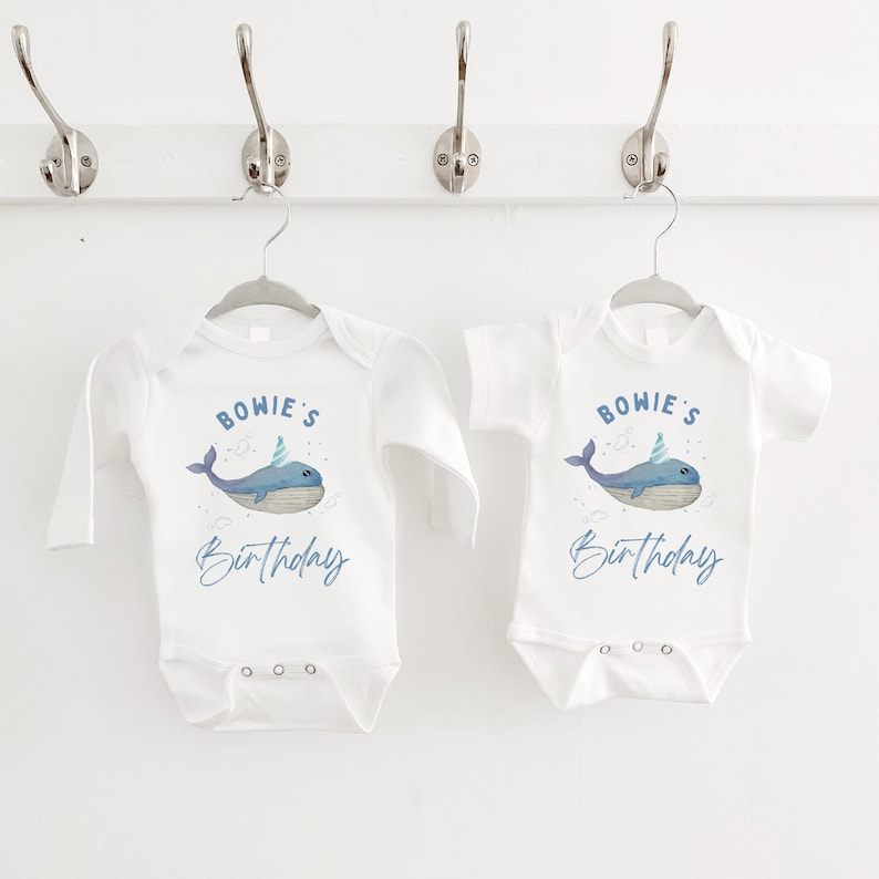 May include: Two white baby onesies with long and short sleeves. The onesies feature a blue whale with a party hat and the text "BOWIE'S Birthday".
