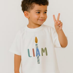 Pode incluir: Uma camiseta branca com o nome "LIAM" em letras coloridas e um desenho de vela. O menino est&aacute; sorrindo e mostrando dois dedos. A camiseta &eacute; feita de um material macio.