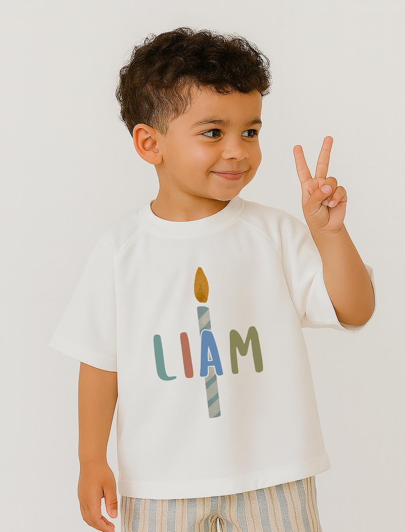 Pode incluir: Uma camiseta branca com o nome "LIAM" em letras coloridas e um desenho de vela. O menino est&aacute; sorrindo e mostrando dois dedos. A camiseta &eacute; feita de um material macio.