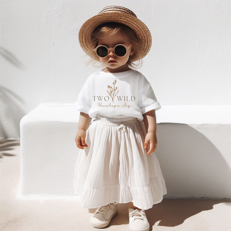 Puede incluir: Un ni&ntilde;o peque&ntilde;o con una camiseta blanca con el texto "TWO WILD Penelope Joy" y un dise&ntilde;o floral. El ni&ntilde;o tambi&eacute;n lleva un sombrero de paja, gafas de sol, una falda blanca y zapatillas blancas.