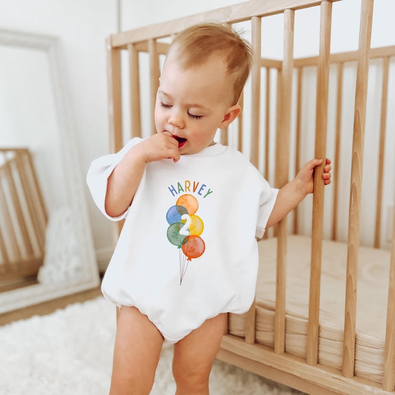 May include: White baby romper with the name "HARVEY" in rainbow letters and a watercolor illustration of balloons. The balloons are in shades of blue, orange, green, and yellow, with the number "2" in the center. The baby is standing near a wooden crib.