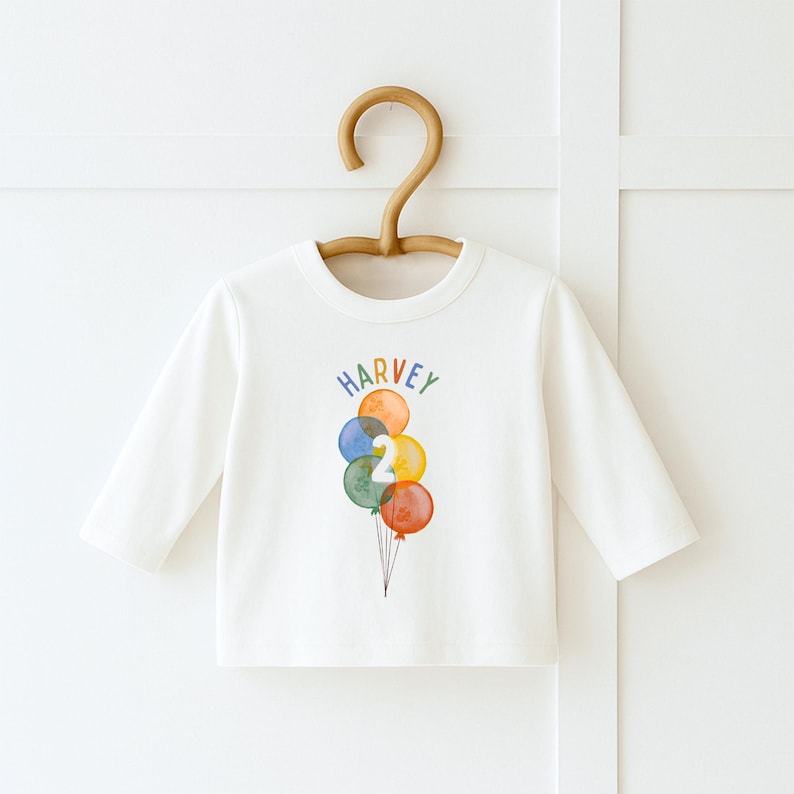 May include: A long-sleeved, off-white shirt with a colorful balloon design. The balloons are in shades of orange, blue, green, and red, with the number "2" and the name "HARVEY" printed on the front. The shirt is hanging on a wooden hanger.