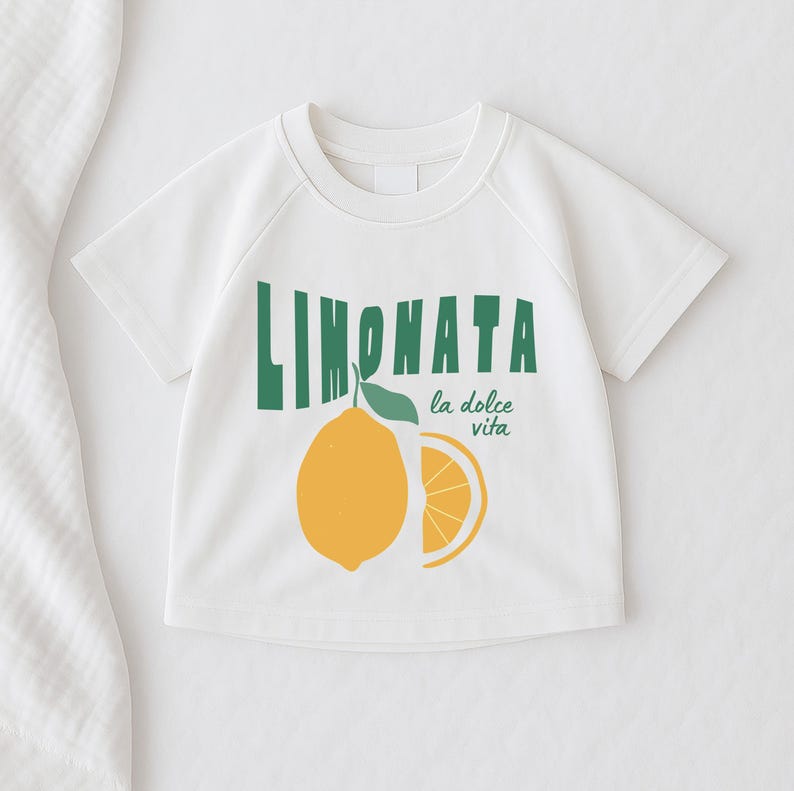May include: White short-sleeved t-shirt with a green and yellow lemon graphic. The shirt features the word "LIMONATA" in green block letters above a yellow lemon illustration and the phrase "la dolce vita" in green.