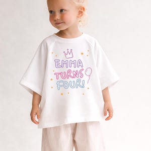 May include: A young child wearing a white t-shirt with the text "EMMA TURNS FOUR!" in colorful letters, a crown, stars, and a balloon graphic. The child is also wearing pink striped pants.