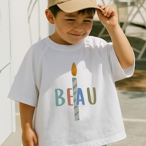 Pode incluir: Camiseta branca com um gr&aacute;fico de vela e o nome "BEAU" em letras coloridas. A vela tem uma chama amarela e um corpo listrado azul e branco. A pessoa est&aacute; usando um bon&eacute; de beisebol bege.