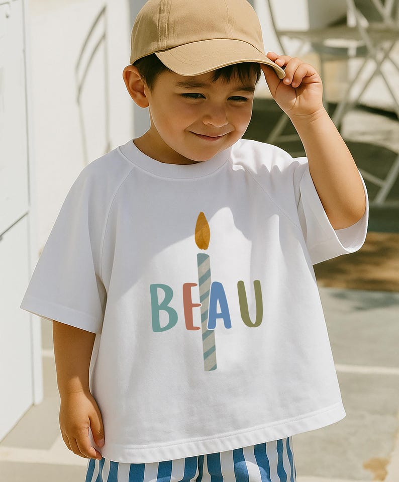 Pode incluir: Camiseta branca com um gr&aacute;fico de vela e o nome "BEAU" em letras coloridas. A vela tem uma chama amarela e um corpo listrado azul e branco. A pessoa est&aacute; usando um bon&eacute; de beisebol bege.