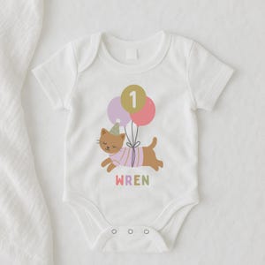 May include: White baby bodysuit with a cartoon cat design. The cat wears a party hat and a striped shirt, holding balloons with the number "1". The name "WREN" is printed in colorful letters below the cat. A cute outfit for a first birthday.