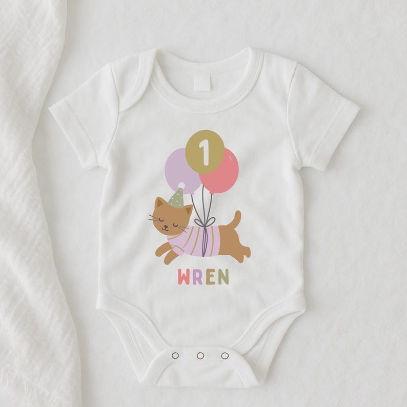 May include: White baby bodysuit with a cartoon cat design. The cat wears a party hat and a striped shirt, holding balloons with the number "1". The name "WREN" is printed in colorful letters below the cat. A cute outfit for a first birthday.