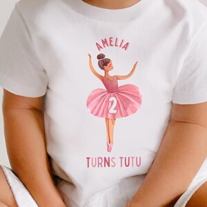 May include: White t-shirt with a pink ballerina graphic and the text "Amelia Turns Tutu" and the number "2".