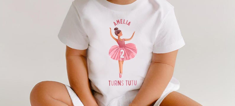 May include: White t-shirt with a pink ballerina graphic and the text "Amelia Turns Tutu" and the number "2".