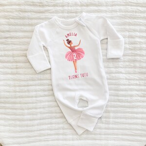 May include: White long-sleeve baby onesie with a pink ballerina illustration and the text "Amelia Turns Tutu 2".