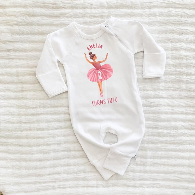 May include: White long-sleeve baby onesie with a pink ballerina illustration and the text "Amelia Turns Tutu 2".