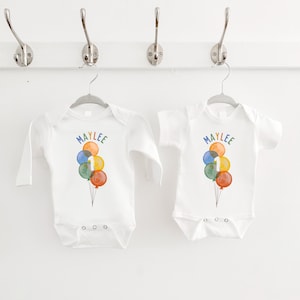 May include: Two white baby onesies, one long-sleeved and one short-sleeved, hanging on a white wall. Each onesie features a colorful watercolor design of balloons with the name "MAYLEE" and the number "1".