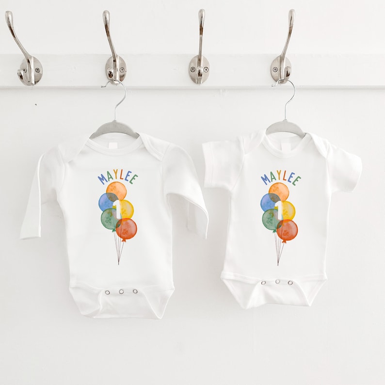 May include: Two white baby onesies, one long-sleeved and one short-sleeved, hanging on a white wall. Each onesie features a colorful watercolor design of balloons with the name "MAYLEE" and the number "1".