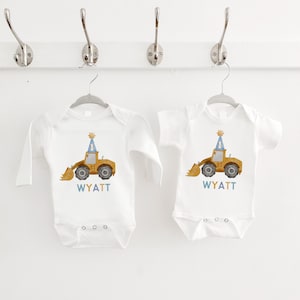 May include: Two white long-sleeved onesies with a yellow construction vehicle wearing a blue party hat. The onesies have the name "WYATT" printed on them.
