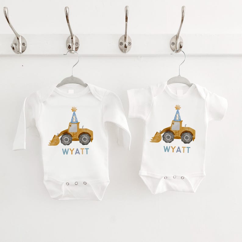 May include: Two white long-sleeved onesies with a yellow construction vehicle wearing a blue party hat. The onesies have the name "WYATT" printed on them.