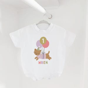 May include: White baby bodysuit with a colorful illustration of a brown cat wearing a party hat and a striped shirt, holding balloons with the number 1. The name "WREN" is printed below the cat.