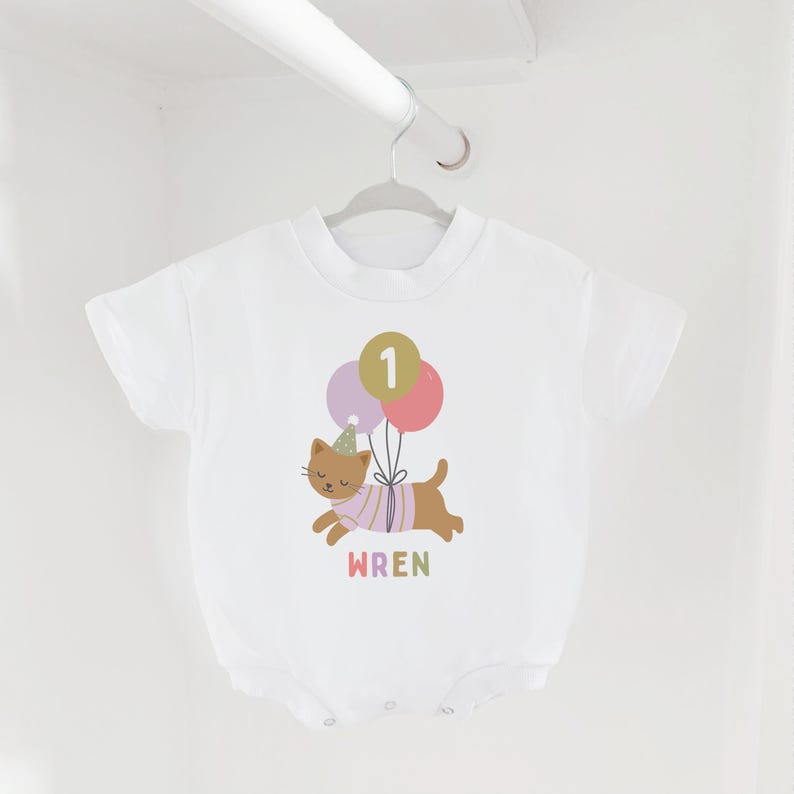 May include: White baby bodysuit with a colorful illustration of a brown cat wearing a party hat and a striped shirt, holding balloons with the number 1. The name "WREN" is printed below the cat.