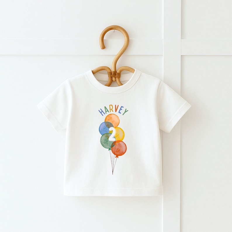 May include: White t-shirt with the name "Harvey" in rainbow letters above a watercolor illustration of colorful balloons. The number "2" is on one of the balloons. The shirt is hanging on a wooden hanger.