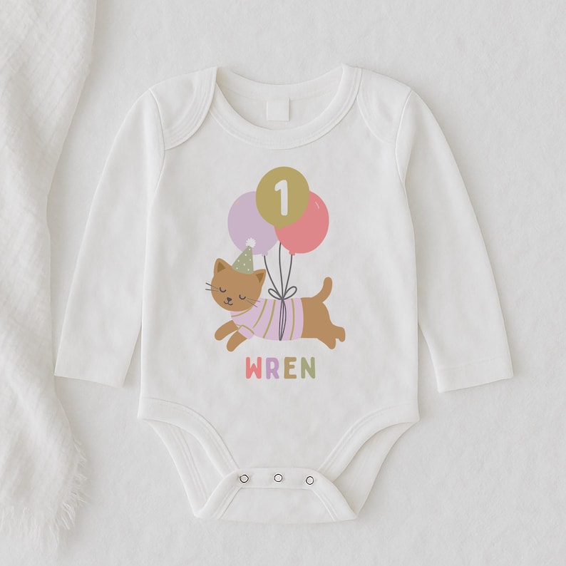 May include: White long-sleeve baby bodysuit with a colorful graphic of a brown cat wearing a party hat and holding balloons. The number "1" and the name "WREN" are also printed on the bodysuit.