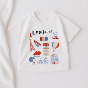 Op de afbeelding: Wit T-shirt met korte mouwen en een kleurrijke Franse afbeelding. Het ontwerp bevat de Eiffeltoren, een baguette, macarons, een koksmuts, een snor en het woord "Bonjour!"