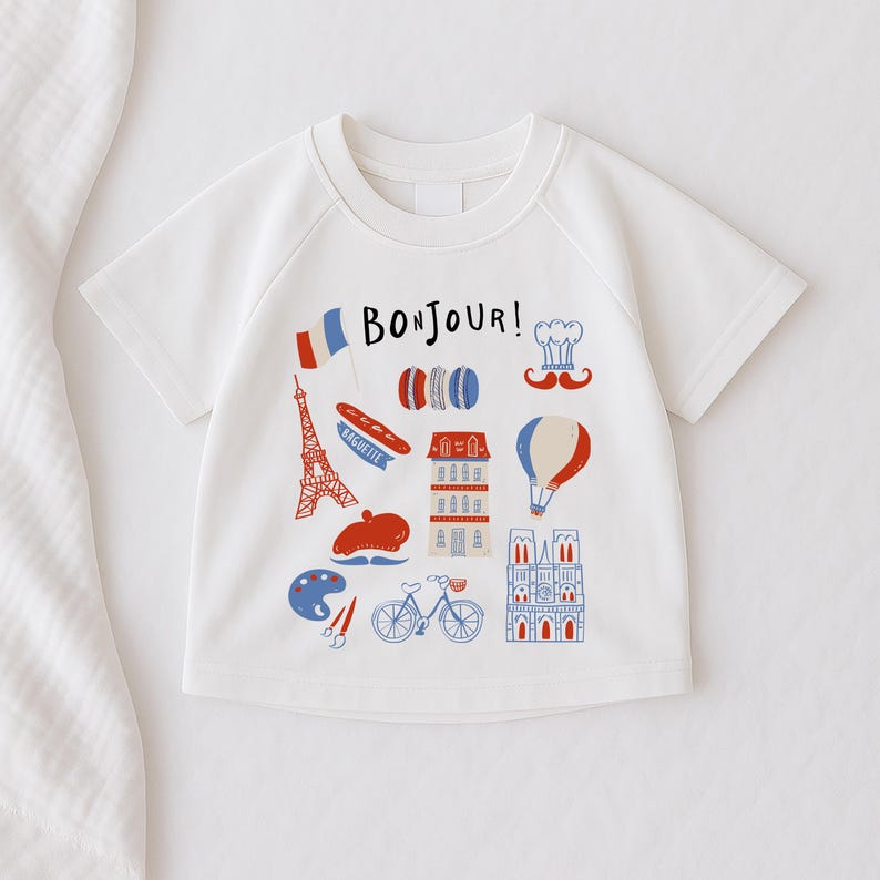 Op de afbeelding: Wit T-shirt met korte mouwen en een kleurrijke Franse afbeelding. Het ontwerp bevat de Eiffeltoren, een baguette, macarons, een koksmuts, een snor en het woord "Bonjour!"