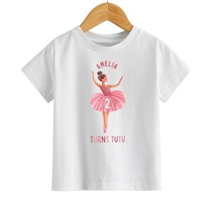 May include: White t-shirt with a pink ballerina graphic and the text "Amelia Turns Tutu" with the number 2 on the ballerina's skirt.